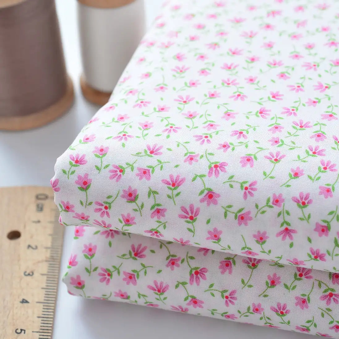 Deux morceaux pliés de Tissu linon de coton fleuri au mètre de Longan Craft France, avec un motif floral rose et vert, sont présentés à côté d'une règle en bois et de deux bobines de fil (marron et blanc) sur une surface blanche.