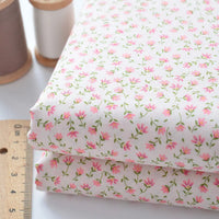Deux pièces pliées de Tissu linon de coton fleuri de Longan Craft France, présentant un motif floral rose, sont empilées à côté d'une règle et de deux bobines de fil, l'une marron, l'autre blanche. Un voile de coton léger idéal pour la confection ou la doublure.