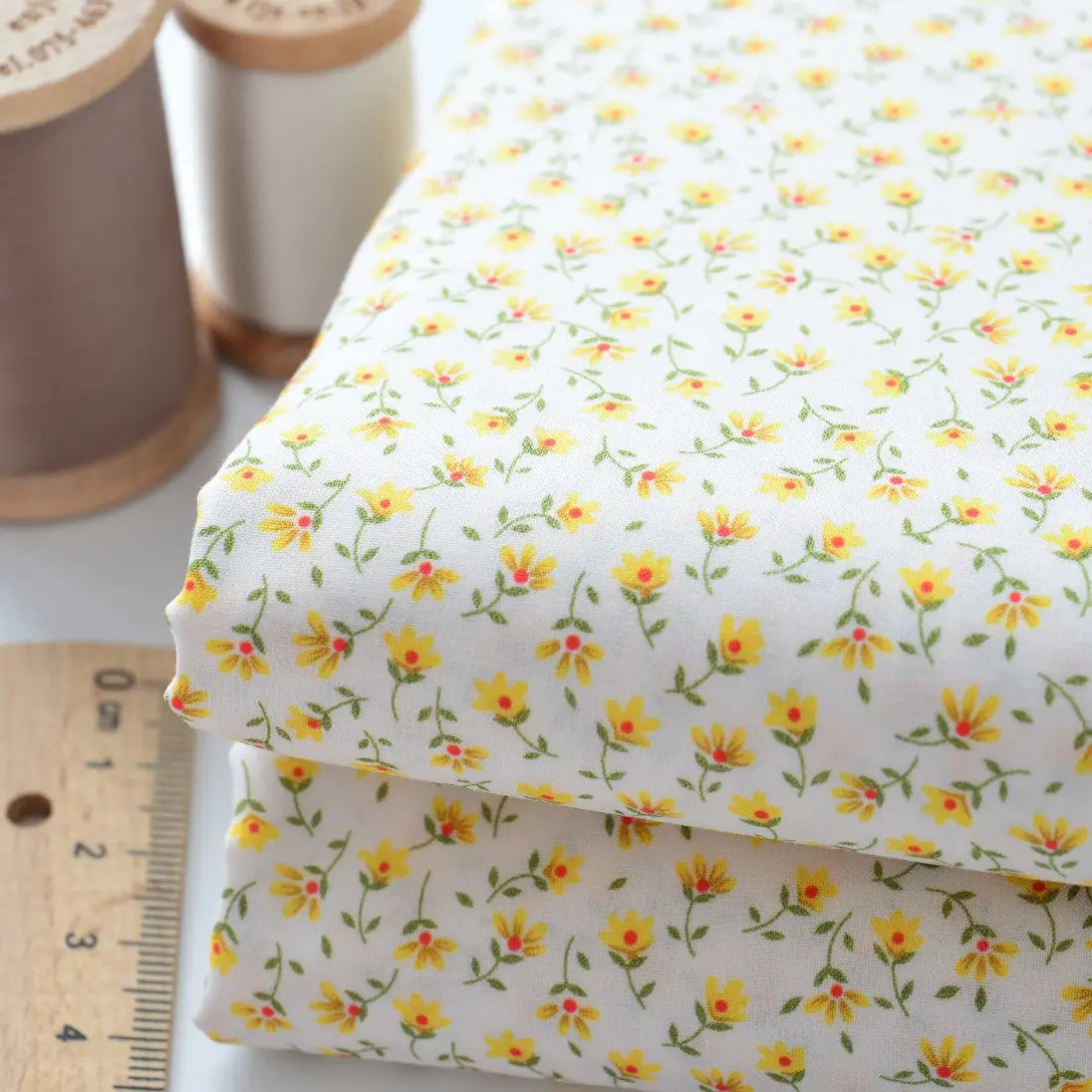 Deux pièces pliées de Tissu linon de coton fleuri au mètre de Longan Craft France, présentant un voile de coton floral jaune, sont présentées à côté d'une règle et de deux bobines de fil (beige et blanc).