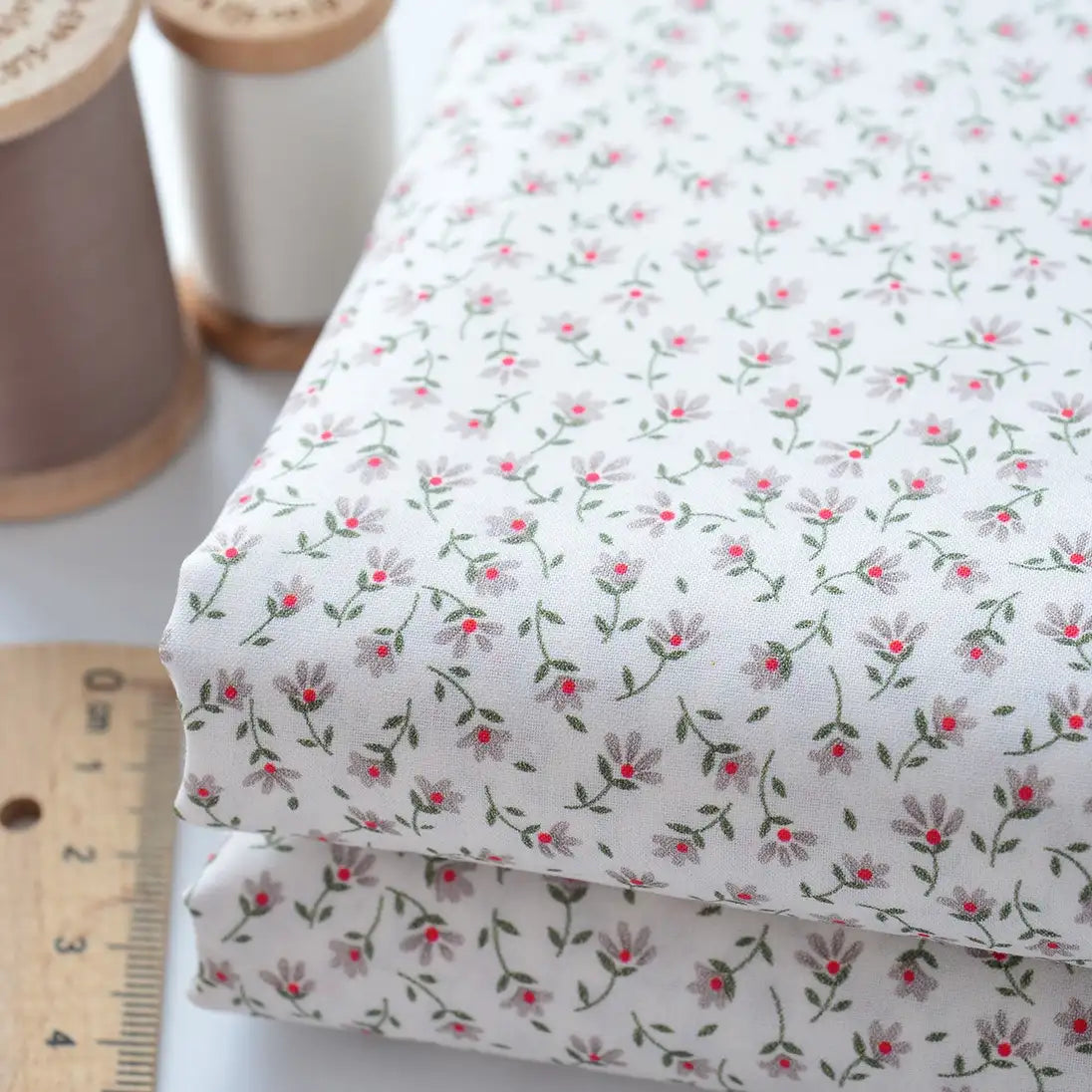 Deux pièces pliées de Tissu linon de coton fleuri de Longan Craft France, présentant un léger imprimé floral, sont présentées à côté d'une règle en bois et de deux bobines de fil sur une surface blanche.