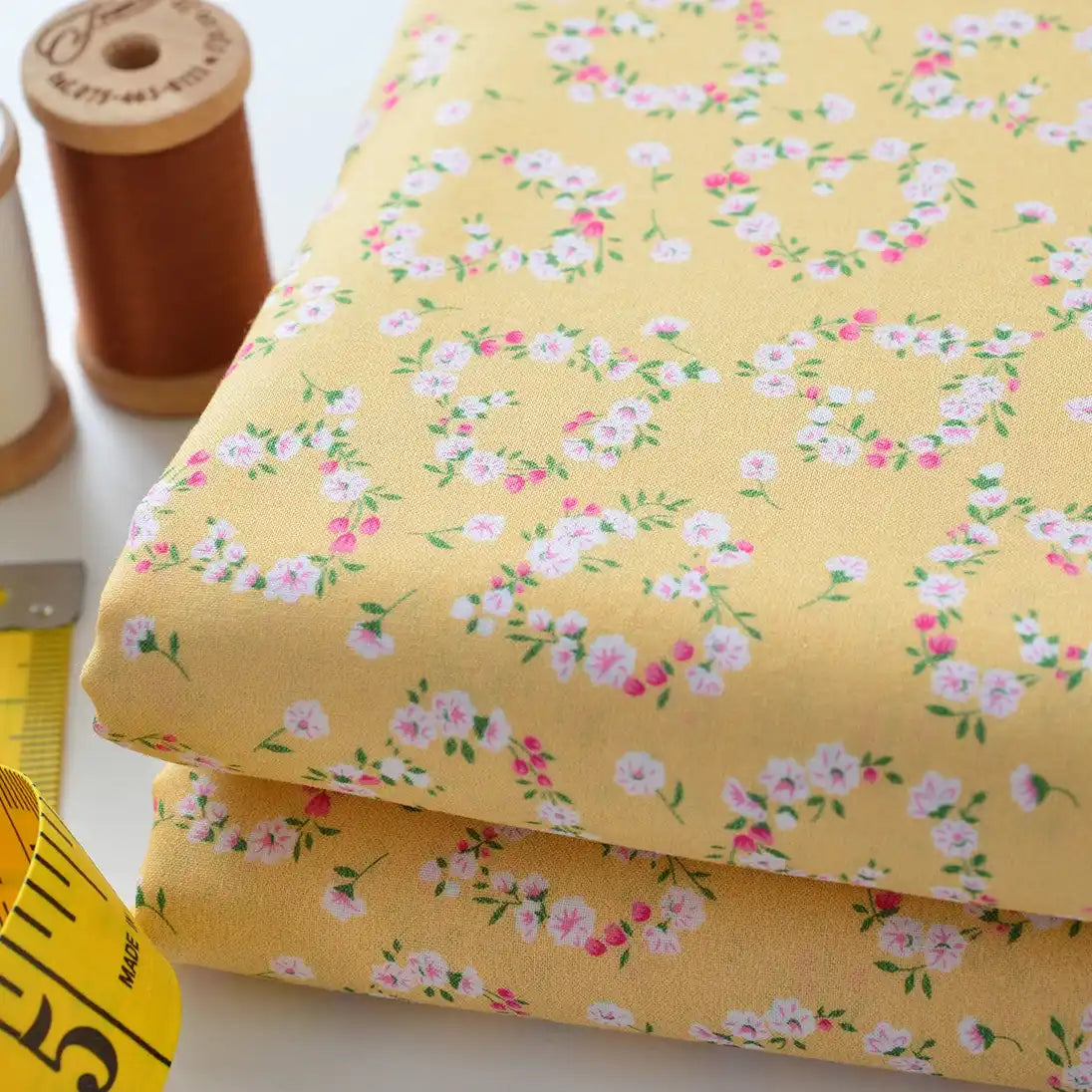 Deux morceaux pliés de Tissu linon de coton fleuri au mètre de Longan Craft France, avec de petits motifs floraux roses et blancs, sont placés à côté de bobines de fil brunes et blanches et d'un mètre ruban jaune sur une surface blanche.