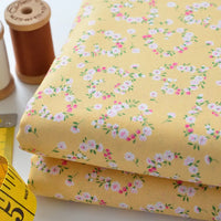 Deux morceaux pliés de Tissu linon de coton fleuri au mètre de Longan Craft France, avec de petits motifs floraux roses et blancs, sont placés à côté de bobines de fil brunes et blanches et d'un mètre ruban jaune sur une surface blanche.