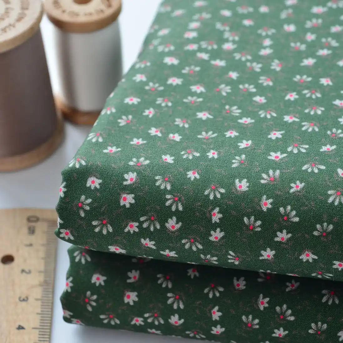 Gros plan sur le Tissu linon de coton fleuri au mètre de Longan Craft France, composé de voile de coton léger vert avec des motifs floraux blancs et roses, présenté avec des bobines de fil en bois et une règle.