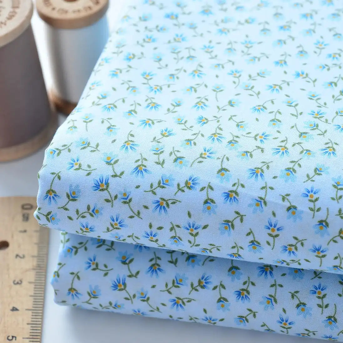 Deux pièces pliées de Tissu linon de coton fleuri au mètre par Longan Craft France, voile de coton bleu clair avec un petit imprimé floral bleu et jaune, présentées à côté d'une règle en bois et de bobines de fil à coudre.