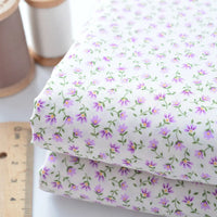 Deux pièces de "Tissu linon de coton fleuri au mètre" de Longan Craft France, présentant un petit motif floral violet, sont empilées à côté d'une règle en bois et de deux bobines de fil. Voile de coton léger idéal pour la confection et la doublure.