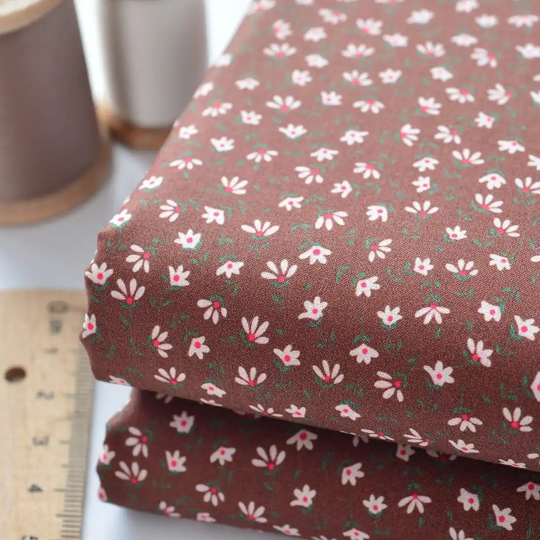 Deux pièces de "Tissu linon de coton fleuri au mètre" de Longan Craft France - un voile de coton léger de couleur marron avec un petit imprimé floral blanc et rose - sont présentées à côté d'une règle en bois et d'une bobine de fil sur une surface blanche.
