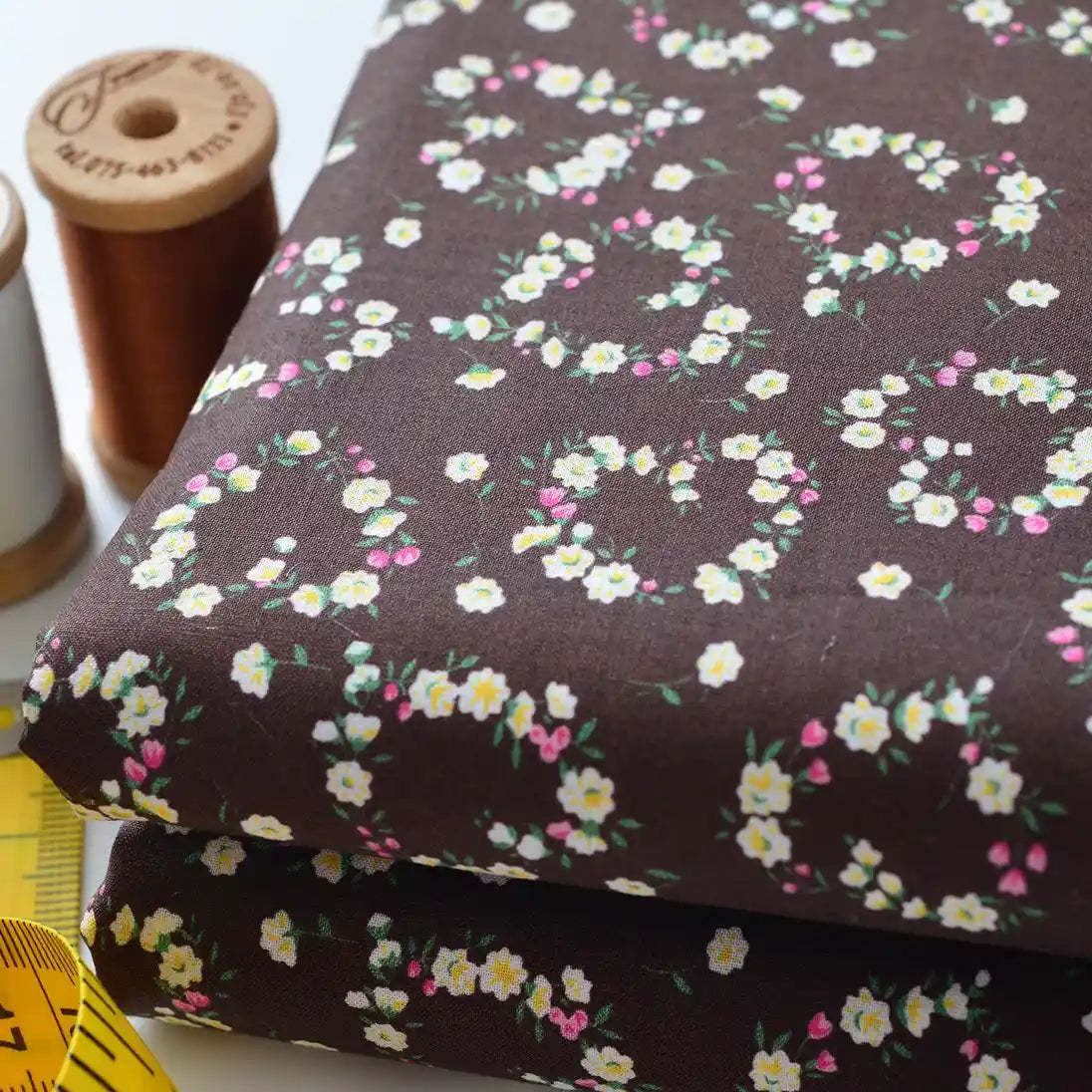 Deux pièces pliées de tissu linon de coton fleuri au mètre de Longan Craft France, brun foncé avec de petits cercles floraux blancs, jaunes et roses, sont présentées avec deux bobines de fil brun et un ruban à mesurer jaune.