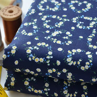 Gros plan sur le tissu linon de coton fleuri au mètre plié de Longan Craft France, un voile de coton léger bleu marine avec de petites fleurs blanches et jaunes, présenté avec des bobines de fil marron et un mètre ruban jaune.