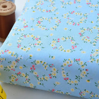 Gros plan sur le Tissu linon de coton fleuri de Longan Craft France : voile de coton bleu clair avec de petites couronnes florales jaunes, roses et vertes. Une bobine de fil marron apparaît partiellement dans le coin supérieur gauche.