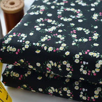 Deux pièces pliées de Tissu linon de coton fleuri au mètre de Longan Craft France présentent un délicat motif floral blanc, jaune et rose. Une bobine de fil marron et un mètre jaune sont partiellement visibles à côté du voile de coton léger.