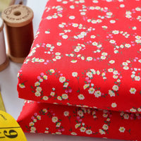 Deux morceaux pliés de Tissu linon de coton fleuri au mètre de Longan Craft France, présentant un motif floral rouge, sont placés à côté de bobines de fil marron et d'un mètre ruban jaune.
