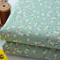 Deux pièces pliées de "Tissu linon de coton fleuri au mètre" de Longan Craft France en vert clair avec de petites fleurs blanches, jaunes et roses. Une bobine de fil marron en bois et un mètre ruban jaune se trouvent à l'arrière-plan.