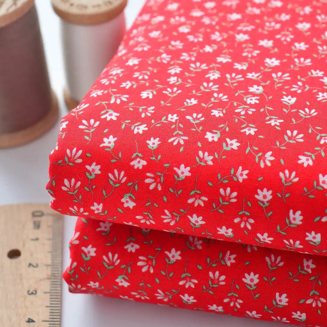 Deux pièces pliées de "Tissu linon de coton fleuri au mètre" de Longan Craft France, comportant un tissu rouge avec un petit motif floral blanc, sont présentées à côté d'une règle en bois et de deux bobines de fil (marron et blanc).