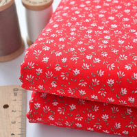 Deux pièces pliées de "Tissu linon de coton fleuri au mètre" de Longan Craft France, comportant un tissu rouge avec un petit motif floral blanc, sont présentées à côté d'une règle en bois et de deux bobines de fil (marron et blanc).