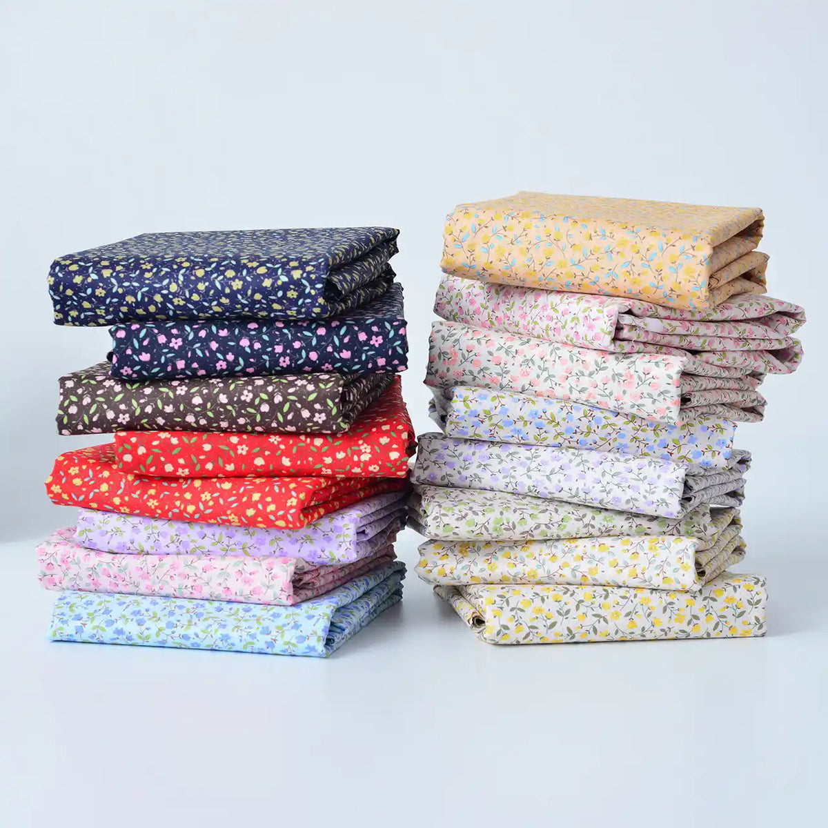 Deux piles bien rangées de Tissu linon de coton fleuri au mètre de Longan Craft France, comprenant un voile de coton léger avec de petits motifs floraux en bleu, rouge, rose, violet, marron, jaune et blanc sur un fond blanc uni.