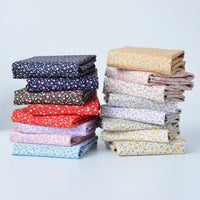 Deux piles bien rangées de Tissu linon de coton fleuri au mètre de Longan Craft France, comprenant un voile de coton léger avec de petits motifs floraux en bleu, rouge, rose, violet, marron, jaune et blanc sur un fond blanc uni.