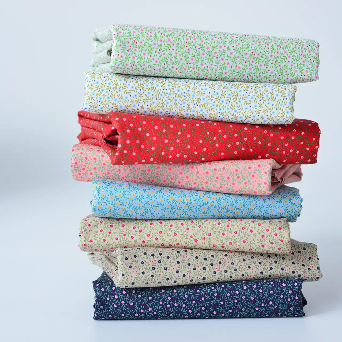 Une pile de Tissu linon de coton fleuri au mètre de Longan Craft France, un voile de coton léger pour les vêtements et les doublures, dans des couleurs assorties et des imprimés floraux, soigneusement disposés sur une surface blanche.