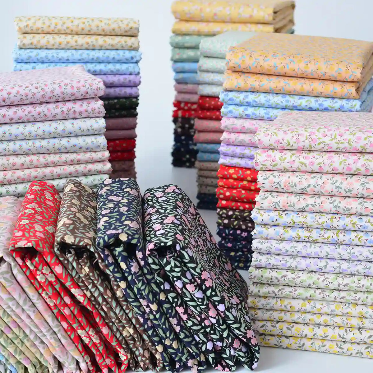 Des piles de tissu linon de coton fleuri de Longan Craft France, un voile de coton léger vendu au mètre pour l'habillement et la doublure, sont soigneusement disposées en rangées dans les couleurs rouge, bleu, vert, jaune, rose et violet avec de petits motifs floraux répétitifs.