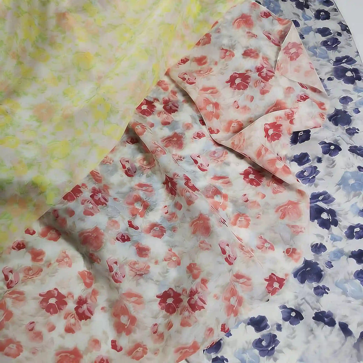 Trois pièces de Longan Craft France Tissu mousseline de soie à imprimé floral au mètre sont disposées : une avec des fleurs jaunes, une avec des fleurs rouges et une avec des fleurs bleues sur fond blanc. Il s'agit d'un tissu léger en soie et coton destiné à la confection de vêtements.
