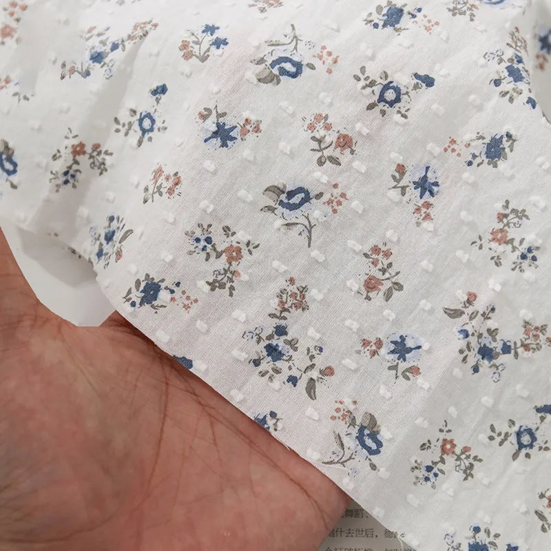 Une main tient le Tissu plumetis à imprimé floral au mètre de Longan Craft France, un tissu plumetis coton respirant orné de fleurs bleues, roses et grises.