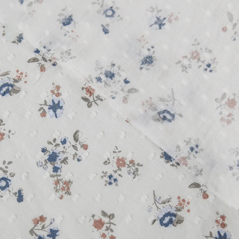 Gros plan sur le tissu plumetis à imprimé floral au mètre en blanc de Longan Craft, qui présente un plumetis de coton léger avec une texture légèrement transparente et des motifs floraux bleus, rouges et bruns dispersés.