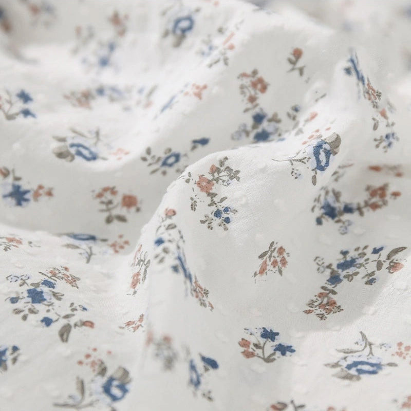 Gros plan sur le Tissu plumetis à imprimé floral au mètre de Longan Craft France, montrant un coton blanc doux à la texture subtilement froissée et de délicats motifs floraux bleus, roses et gris éparpillés sur la surface.