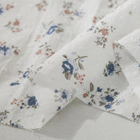 Gros plan sur le tissu plumetis à imprimé floral au mètre de Longan Craft France, un tissu de coton respirant et légèrement transparent avec des imprimés floraux bleus, gris et brun-rouge épars, présentés en couches doucement pliées.