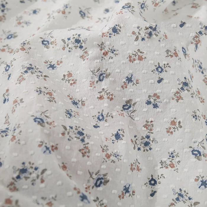 Le tissu plumetis à imprimé floral au mètre de Longan Craft France est un coton respirant à la texture subtilement pointillée et à l'imprimé floral délicat bleu, rose et beige avec des feuilles vertes. Le tissu est légèrement froissé.