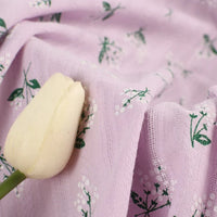 Une tulipe de couleur crème repose sur du Tissu dobby imprimé floral au mètre de Longan Craft France, un tissu de coton violet clair avec de petites tiges vertes et des fleurs blanches, dont la surface est légèrement texturée et les plis souples.