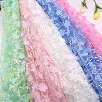 Plusieurs rouleaux de Tissu sequin floral au mètre -résille à sequins en relief et tissu brodé scintillant- de Longan Craft France sont présentés en vert, rose, blanc et bleu avec des sequins étincelants et des applications florales en 3D. Deux fleurs jaunes se trouvent à proximité.