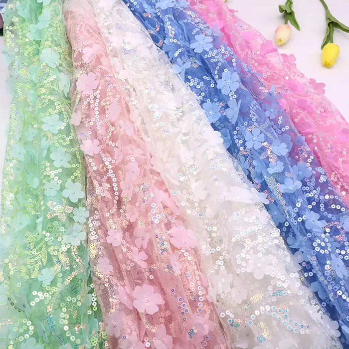 Plusieurs rouleaux de Tissu sequin floral au mètre -résille à sequins en relief et tissu brodé scintillant- de Longan Craft France sont présentés en vert, rose, blanc et bleu avec des sequins étincelants et des applications florales en 3D. Deux fleurs jaunes se trouvent à proximité.