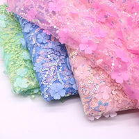 Trois tissus pliés de Longan Craft France - Tissu sequin floral au mètre, Tissu résille à sequins en relief et Tissu brodé scintillant - brillent côte à côte en vert, bleu et rose avec des sequins et des applications florales en 3D sur une surface blanche.