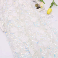 Le Longan Craft France Tissu sequin floral au mètre, une résille blanche à sequins en relief, est présenté avec une broderie florale irisée. Des tulipes jaunes et une partie d'un magazine apparaissent dans le coin supérieur droit.