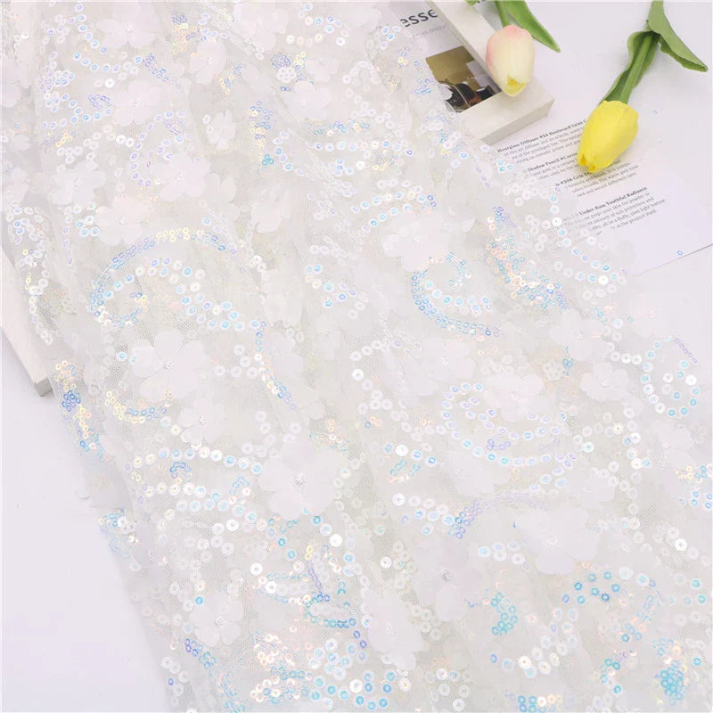 Le Longan Craft France Tissu sequin floral au mètre, une résille blanche à sequins en relief, est présenté avec une broderie florale irisée. Des tulipes jaunes et une partie d'un magazine apparaissent dans le coin supérieur droit.