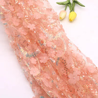 Le Tissu sequin floral au mètre de Longan Craft France, une résille à sequins brodée en relief de couleur pêche, est présenté à côté de deux tulipes jaunes sur une surface claire.