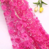 Le "Tissu sequin floral au mètre" de Longan Craft France, une dentelle rose vif avec des applications florales en 3D et des paillettes, est présenté à plat. Deux tulipes jaunes avec des feuilles vertes se trouvent en diagonale à l'arrière-plan.