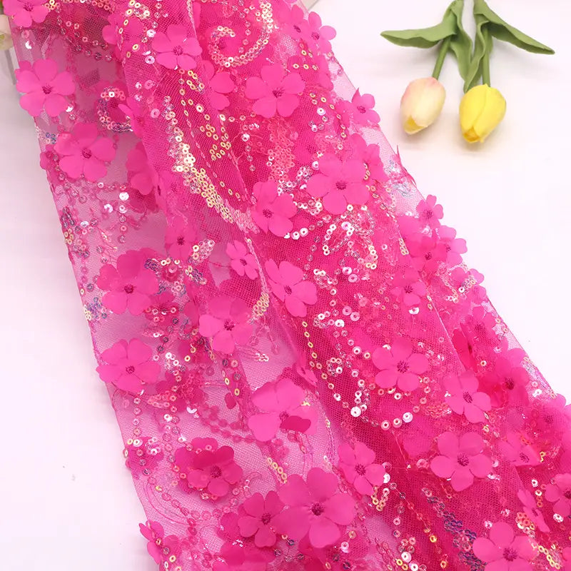 Le "Tissu sequin floral au mètre" de Longan Craft France, une dentelle rose vif avec des applications florales en 3D et des paillettes, est présenté à plat. Deux tulipes jaunes avec des feuilles vertes se trouvent en diagonale à l'arrière-plan.
