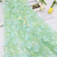 Un "Tissu sequin floral au mètre" vert clair de Longan Craft France, décoré de paillettes et d'applications florales en 3D, est drapé sur une surface blanche avec deux tulipes jaunes et du papier imprimé en arrière-plan.