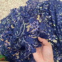 Une main tient le tissu sequin floral au mètre de Longan Craft France, une résille bleu foncé à sequins en relief avec broderie florale en 3D, tandis que d'autres tissus scintillants pour accessoires de robe sont étalés en dessous.