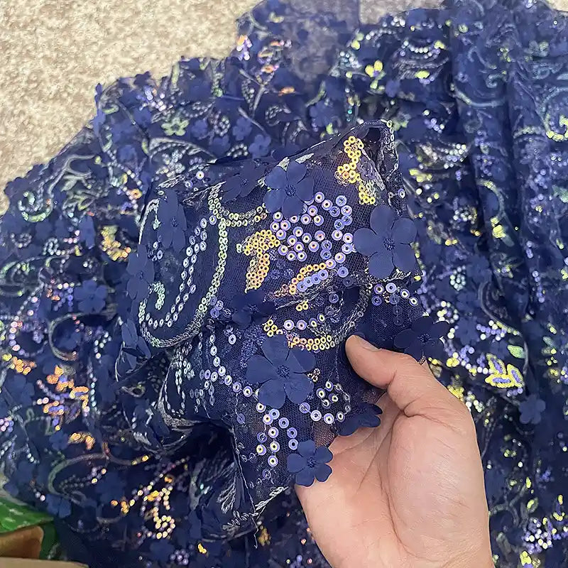 Une main tient le tissu sequin floral au mètre de Longan Craft France, une résille bleu foncé à sequins en relief avec broderie florale en 3D, tandis que d'autres tissus scintillants pour accessoires de robe sont étalés en dessous.