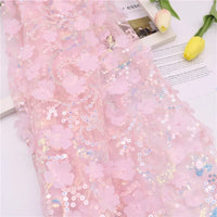 Gros plan sur le Tissu sequin floral au mètre de Longan Craft France, composé d'une résille rose à sequins en relief avec des applications florales en 3D, présenté sur fond blanc avec deux tulipes jaunes et un magazine en arrière-plan.
