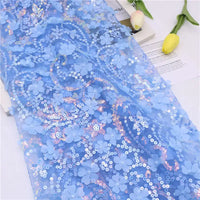 Gros plan sur le Tissu sequin floral au mètre de Longan Craft France, une résille à sequins en relief bleu clair avec des applications florales en 3D et des sequins irisés, drapée sur une surface blanche à côté de tulipes et de papiers jaunes.