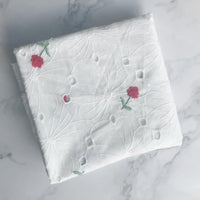 Un morceau de Tissu coton brodé ajouré à fleurs de Longan Craft France, composé de coton blanc avec de délicates broderies florales rouges et vertes, est présenté sur une surface en marbre, idéale pour créer des robes souples.