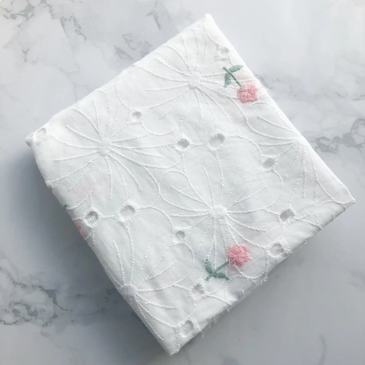 Un morceau du Tissu coton brodé ajouré à fleurs de Longan Craft France, un tissu de coton blanc doux avec broderie florale, avec des fleurs roses et des feuilles vertes, repose sur une surface en marbre gris clair. Idéal pour les robes.