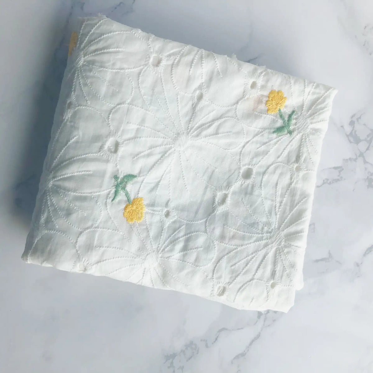 Un morceau plié du tissu coton brodé ajouré à fleurs au mètre de Longan Craft France, un tissu de coton blanc avec une délicate broderie florale, repose sur une surface en marbre clair.