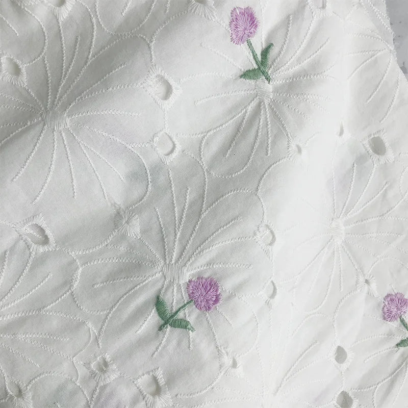 Le tissu coton brodé ajouré à fleurs de Longan Craft France est un coton blanc doux, orné de grandes fleurs blanches et de petites fleurs brodées violettes et vertes, idéal pour robes.