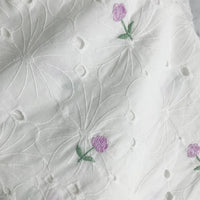 Le tissu coton brodé ajouré à fleurs de Longan Craft France est un coton blanc doux, orné de grandes fleurs blanches et de petites fleurs brodées violettes et vertes, idéal pour robes.