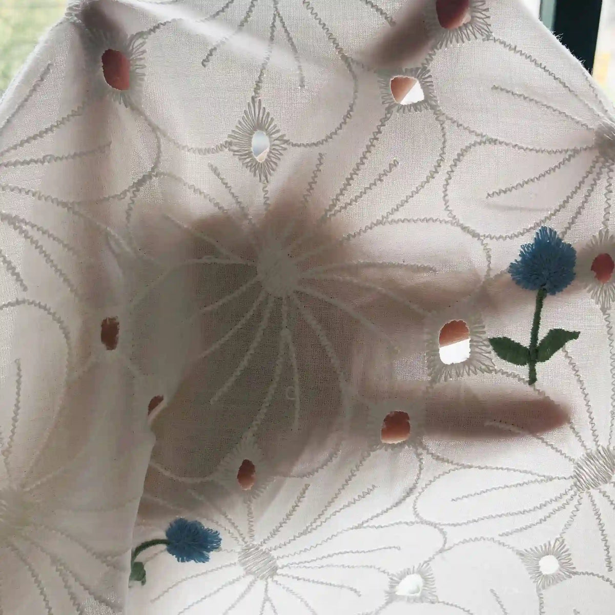 Une main apparaît derrière le tissu coton brodé ajouré à fleurs au mètre de Longan Craft France, un tissu de coton blanc avec des broderies florales et des détails découpés. La lumière passe à travers, révélant les fleurs bleues et le doux contour de la main.