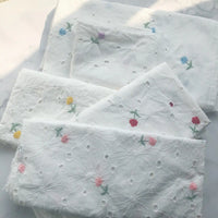 Trois pièces pliées de Tissu coton brodé ajouré à fleurs de Longan Craft France, un tissu de coton blanc doux avec des broderies florales, sont disposées sur une surface légère - idéale pour les robes et disponible au mètre.
