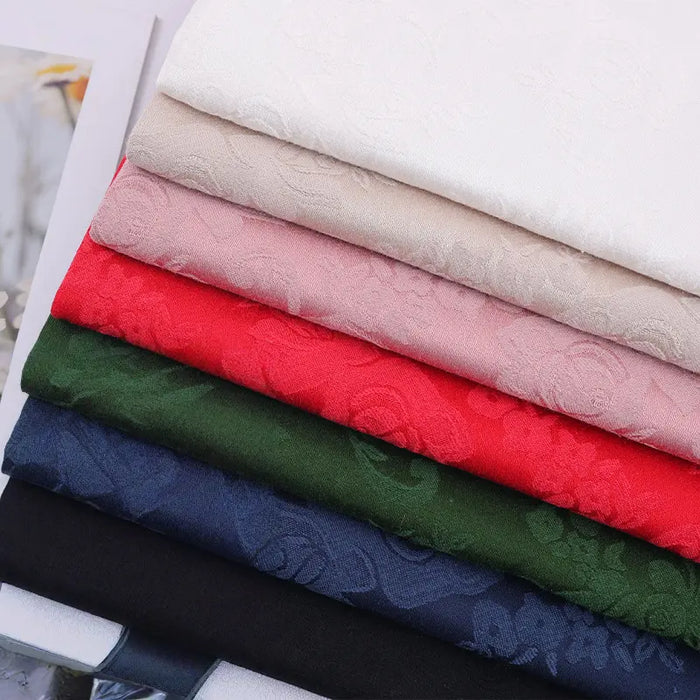 Un ensemble de sept tissus damassés de coton à motif floral de Longan Craft France, en blanc, beige, rose, rouge, vert, bleu marine et noir, empilés avec soin, présente une élégante texture jacquard de chinoiserie, parfaite pour les nappes damassées.
